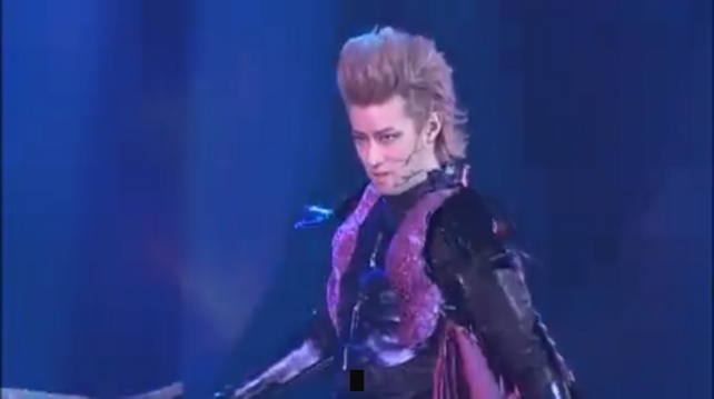 Nelo Angelo wearing a black suit with purple accents, plus his hair is done up