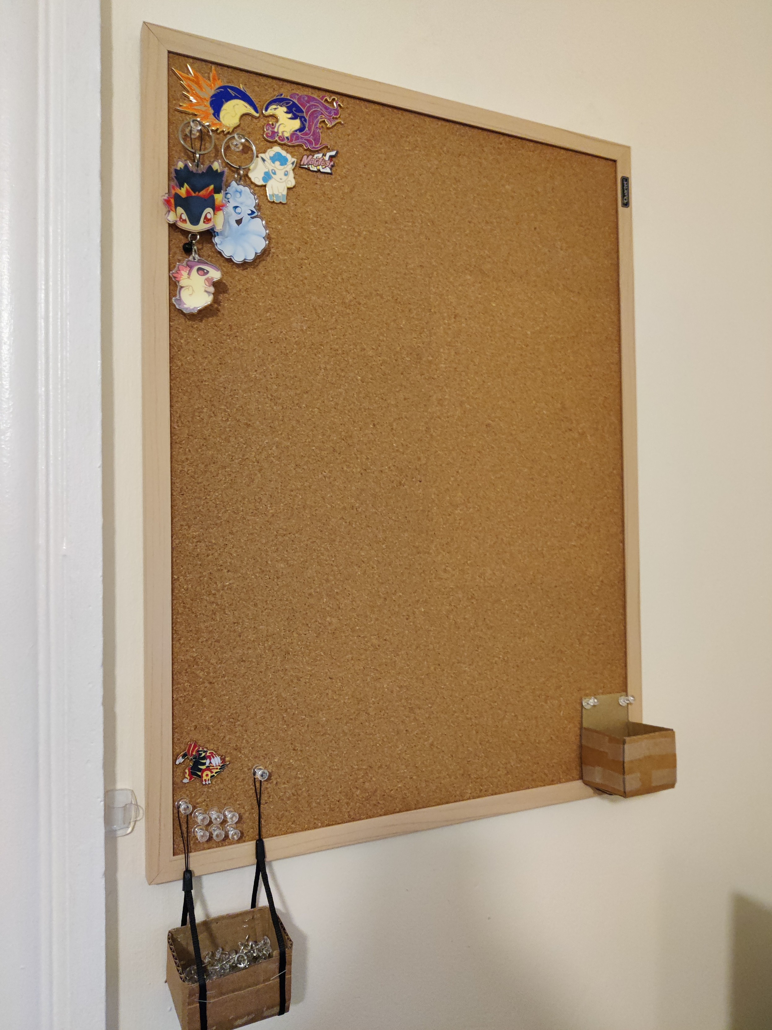 a corkboard hung on the wall. there are various enamel pins and other decorations in the upper left corner. there are small makeshift boxes in the lower right and left corners, one of which is full of pushpins and is being protected by a groudon pin